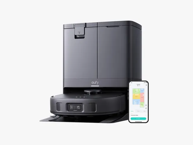Black circular robotic vacuum docked in a tall black station. Small mobile phone to the right, displaying a floorplan.